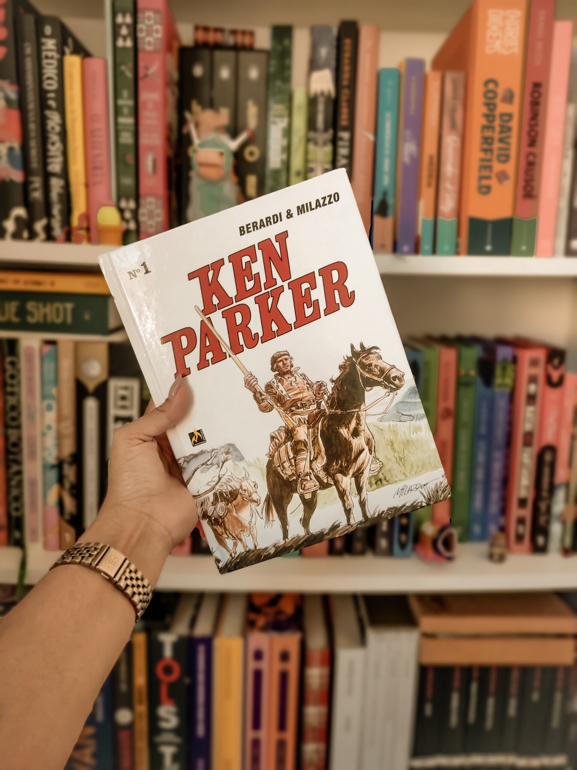 foto da capa da hq Ken Parker, nº 1, publicado pela Mythos Editora no Brasil em 2021, na frente de uma estante de livros; na capa, Ken Parker aparece montando um cavalo e o título da hq está escrito em vermelho.