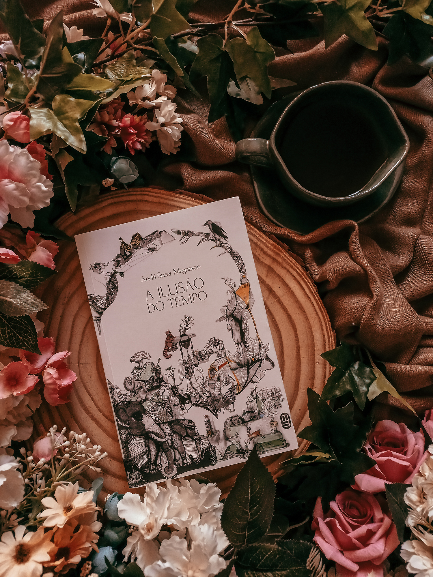 Sobre um tronco de árvore está a edição do livro A Ilusão do Tempo, de Andri Snaer Magnason, publicado pela Editora Morro Branco; ao redor, cobrindo a parte de baixo e esqueurda do cenário, estão várias flores artificiais verdes, brancas, cor de rosa e azuis; do lado direito, no canto superior, em cima de um lenço de cor bege, está uma xícara sobre um pires, na cor verde, imitando o formato de uma folha.