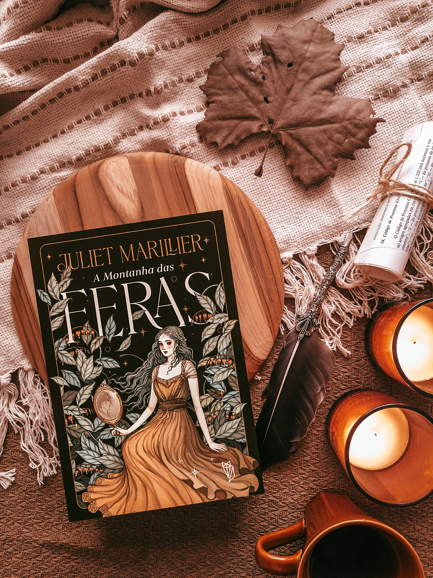 Cenário de livro em flat lay, mostrando a capa de A Montanha das Feras como se já estivesse impresso, ao lado de velas, caneta com pena, papel enrolado, caneca e folha de árvore.