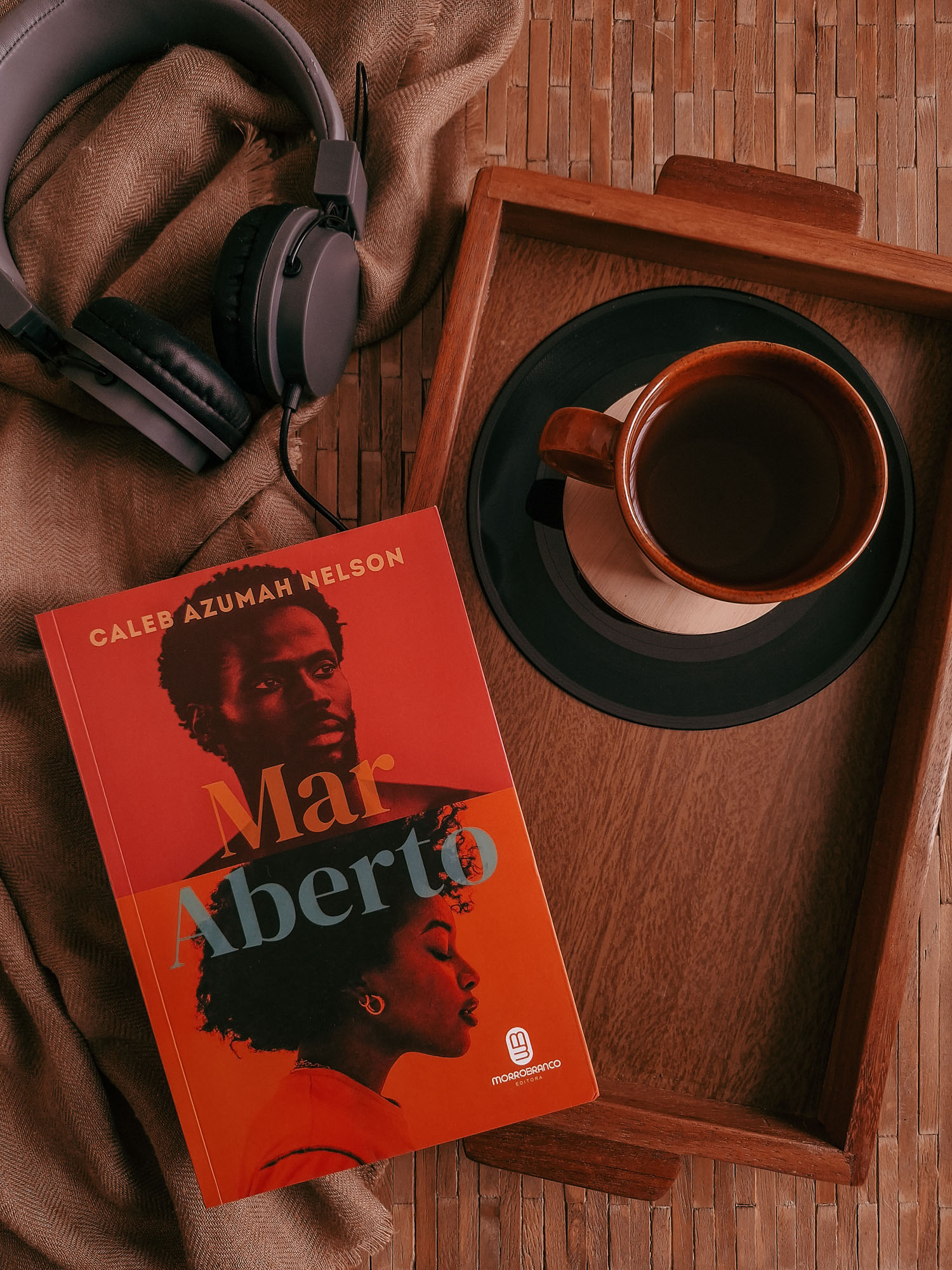 Foto em flat lay com livro Mar Aberto à esquerda e abaixo, por sobre uma bandeja de madeira que cobre a metade direta da foto sobre fundo de madeira, ao lado esquerdo da foto, um lenço cor caramelo com um fone de ouvido estilo headphone acima do livro, à esquerda da foto também.